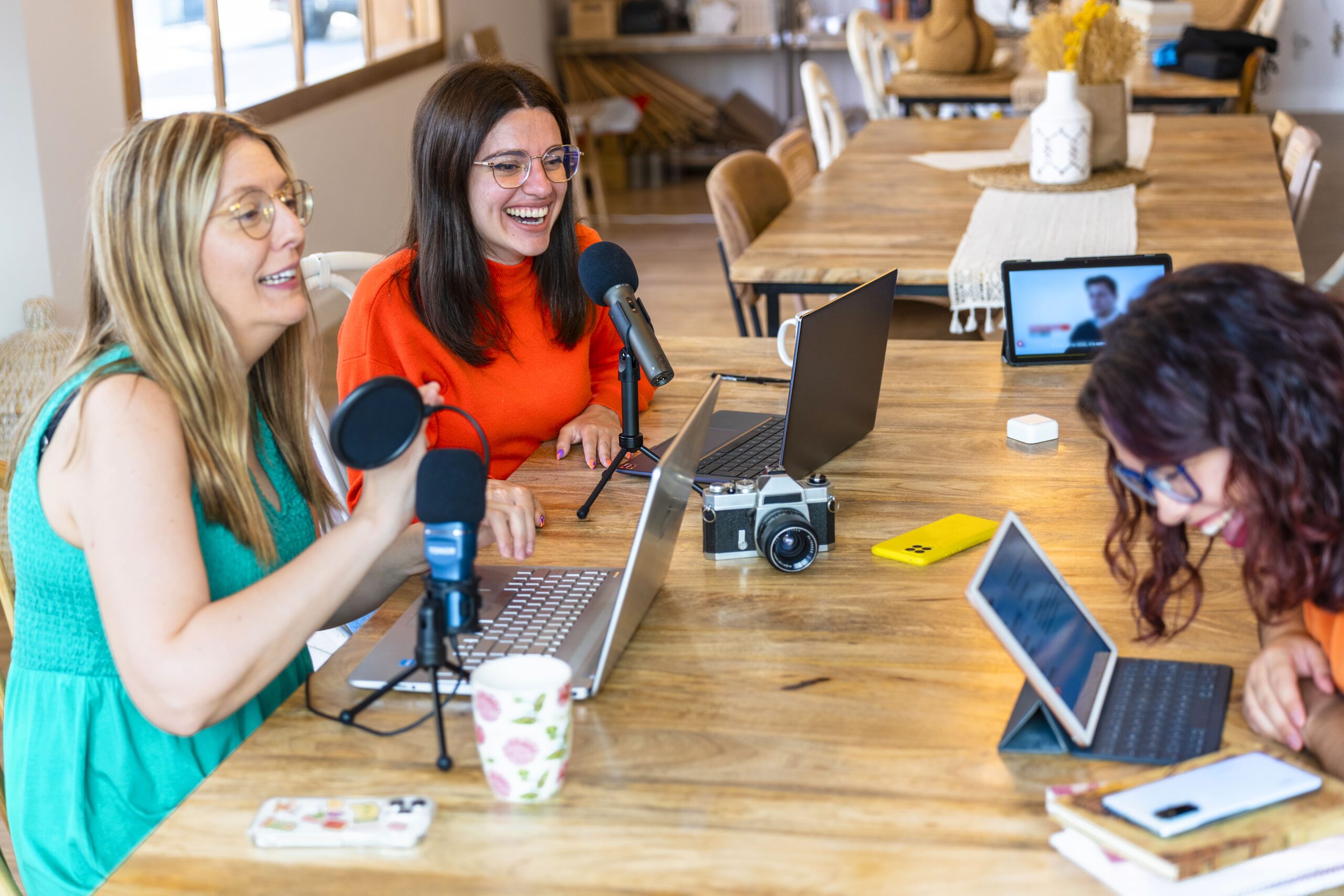 Three women laughing while recording a podcast around a table with microphones and laptops.